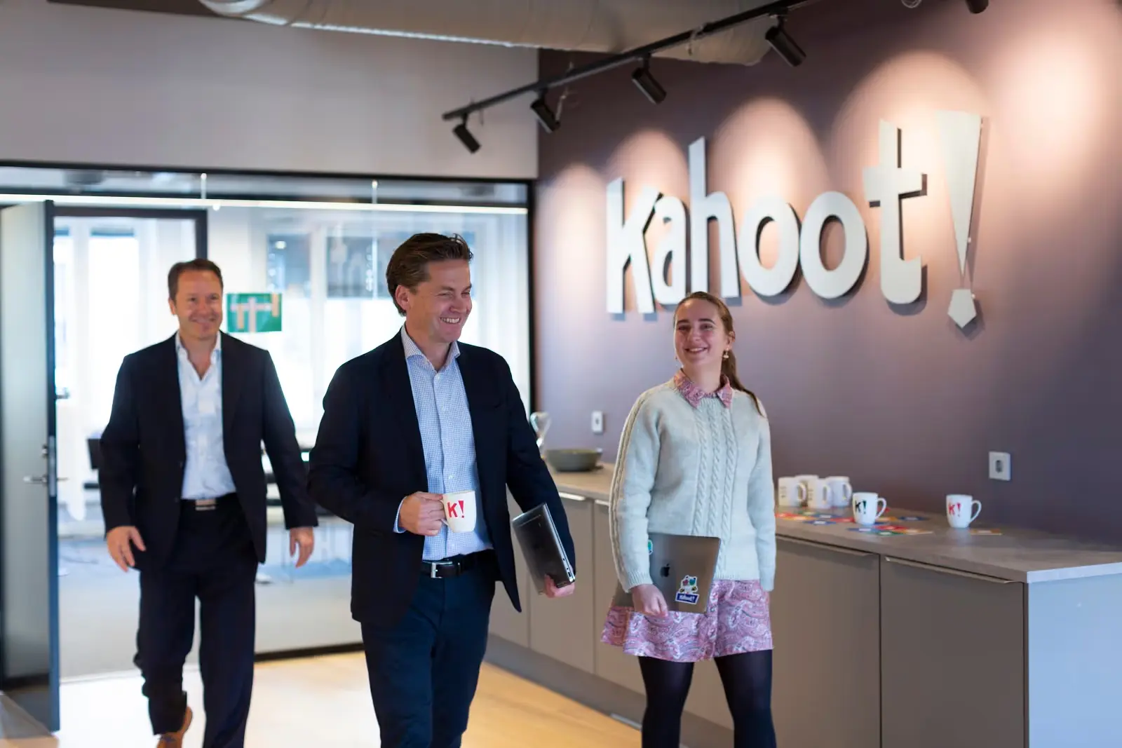 Tre smilende personer går i Kahoot-kontor med laptopper og kopper, logo på veggen bak.